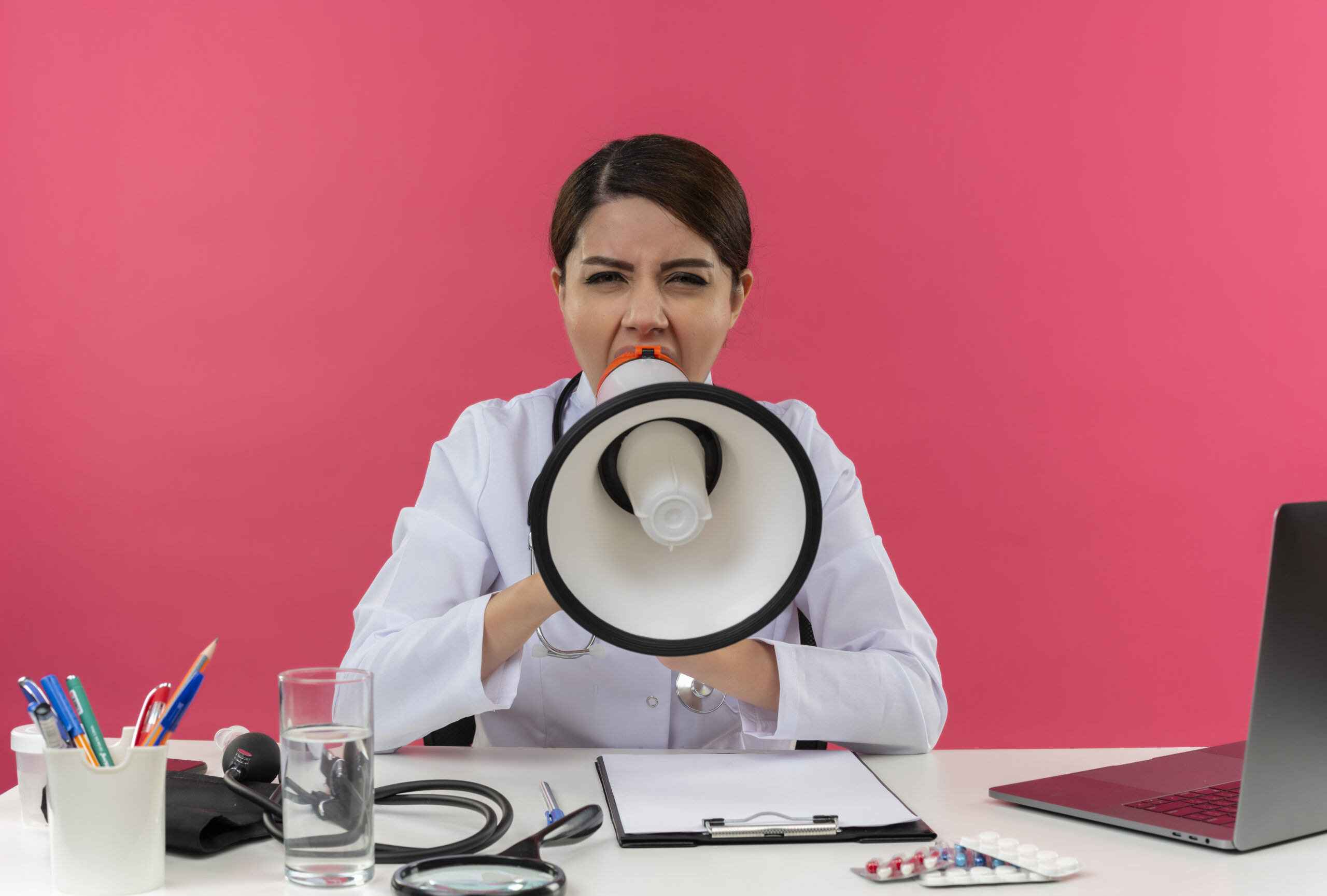 young female doctor wearing medical robe and stethoscope sitting at desk with medical tools and laptop looking at camera shouting in loud speaker isolated on pink background