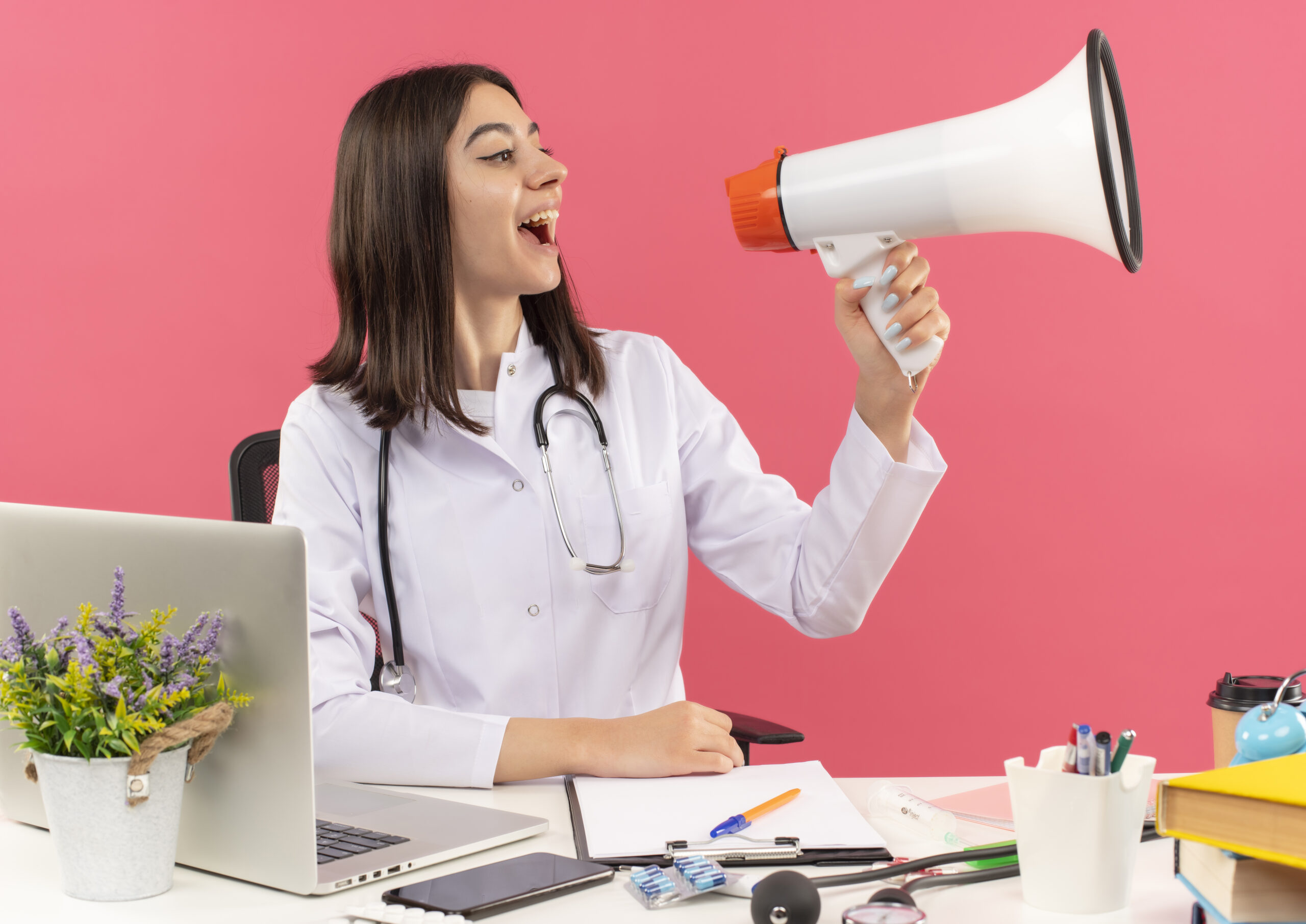 young woman doctor in white coat with stethoscope around her neck shouting to megaphone happy and excited sitting at the table with laptop over pink background